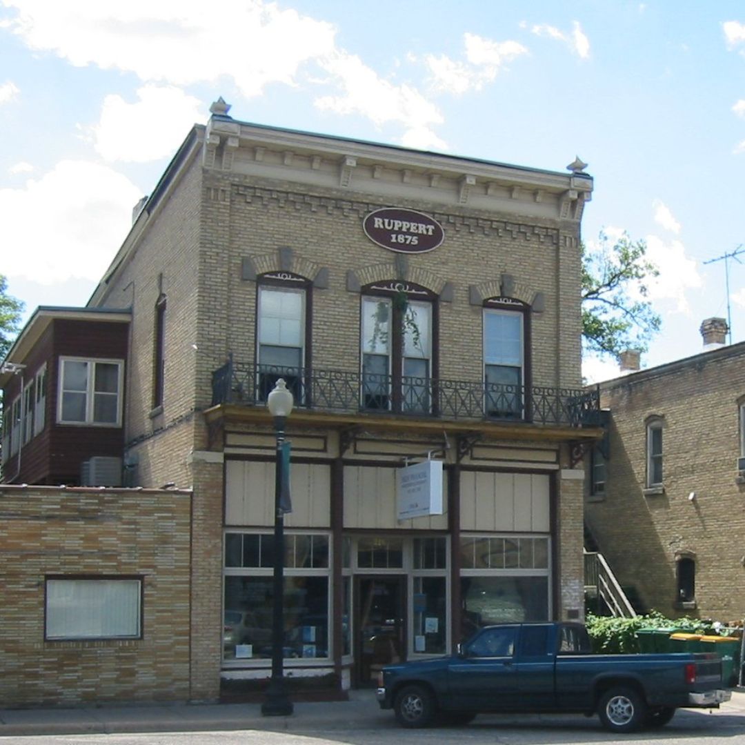 A classic cream-colored brick building from 1875, labeled "Ruppert," characteristic of the architecture in the Jordan, MN Historic District, under a clear sky.